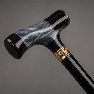 May include: A walking cane featuring a T-shaped handle with a black and white marbled pattern. The handle connects to a smooth black shaft via a decorative gold-colored metal collar with an intricate woven texture pattern around its circumference.