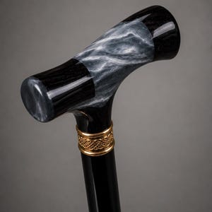 May include: A walking cane featuring a T-shaped handle with a polished black finish and a swirling grey and white marbled pattern. A gold-toned metal collar with an intricate woven knot design sits directly below the handle on the black shaft.