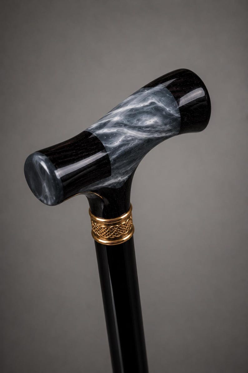 May include: A walking cane featuring a T-shaped handle with a polished black finish and a swirling grey and white marbled pattern. A gold-toned metal collar with an intricate woven knot design sits directly below the handle on the black shaft.