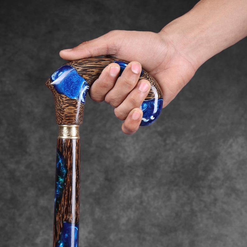 May include: A wooden walking cane with a curved handle. The handle and shaft feature a combination of brown wood and blue resin. The handle is held by a hand. The cane has a gold-colored metal band near the top of the shaft.