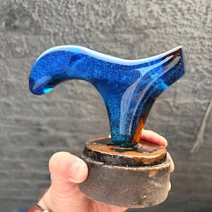 Puede incluir: Un mango de bast&oacute;n artesanal con una parte superior de resina azul vibrante, salpicada de purpurina, y una base de madera. El mango est&aacute; dise&ntilde;ado con una forma curva para un agarre c&oacute;modo. La base es de color marr&oacute;n oscuro.
