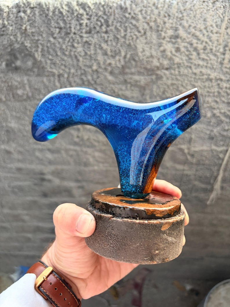 Puede incluir: Un mango de bast&oacute;n artesanal con una parte superior de resina azul vibrante, salpicada de purpurina, y una base de madera. El mango est&aacute; dise&ntilde;ado con una forma curva para un agarre c&oacute;modo. La base es de color marr&oacute;n oscuro.