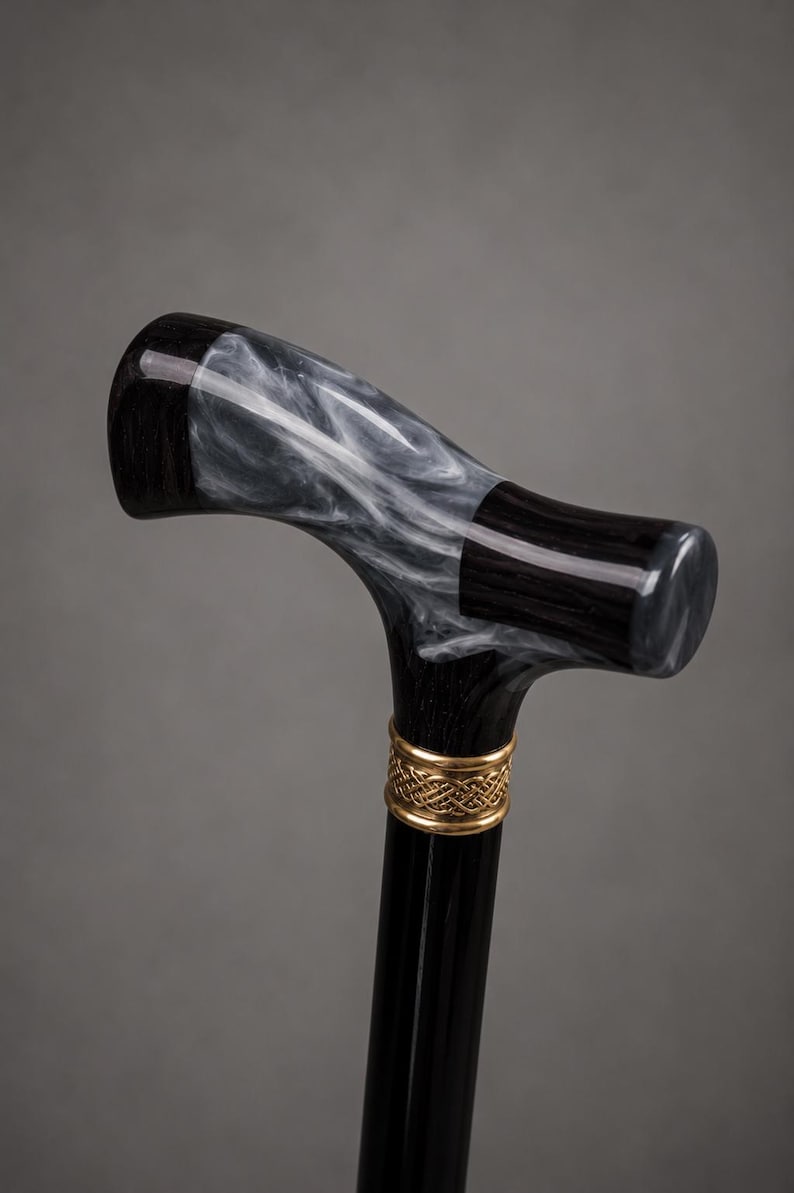 May include: A walking cane featuring a T-shaped handle made of polished black wood and a marbled white and gray resin insert. A gold-colored metal collar with an intricate woven pattern sits just below the handle on the black wooden shaft.