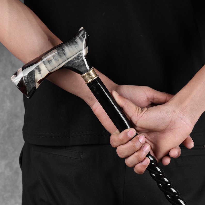 May include: A black and white walking cane with a decorative handle. The handle is made of a light-colored material with dark accents, and the shaft is black with a twisted pattern.  The cane features a metallic band near the handle. This elegant walking stick is a stylish mobility aid.