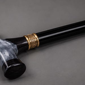May include: A walking cane featuring a T-shaped handle with a marbled black and white resin grip. The shaft is solid black, accented by a decorative gold-toned metal collar with an intricate woven pattern connecting the handle to the main body.