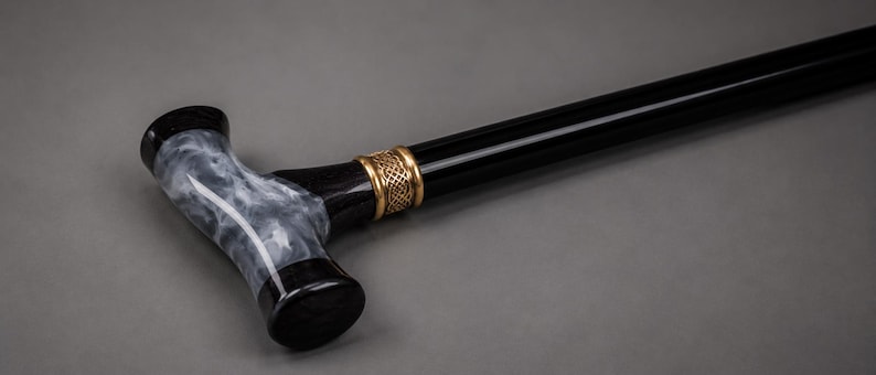 May include: A walking cane featuring a T-shaped handle with a marbled black and white resin grip. The shaft is solid black, accented by a decorative gold-toned metal collar with an intricate woven pattern connecting the handle to the main body.