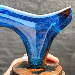 Puede incluir: Un tap&oacute;n de botella artesanal de resina azul con un efecto brillante similar a una galaxia. Montado sobre una base de madera con un acabado oscuro. El dise&ntilde;o presenta una forma curva &uacute;nica.