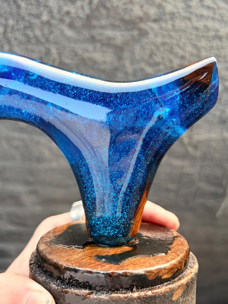 Puede incluir: Un tap&oacute;n de botella artesanal de resina azul con un efecto brillante similar a una galaxia. Montado sobre una base de madera con un acabado oscuro. El dise&ntilde;o presenta una forma curva &uacute;nica.