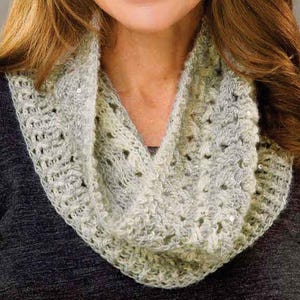 May include: A white and gray crocheted infinity scarf with silver sequins. The scarf is worn by a person with long brown hair.