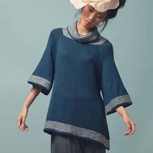 Puede incluir: Un suéter de color verde azulado con cuello alto y mangas anchas. El suéter tiene un ribete gris y blanco estampado en el cuello, las mangas y el dobladillo. La modelo lleva un sombrero color crema.