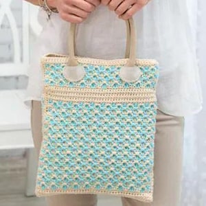 May include: A crocheted tote bag with a light blue and cream-colored pattern. The bag has cream-colored handles and trim. The bag is held by a person wearing a white shirt and tan pants. The bag is rectangular in shape.