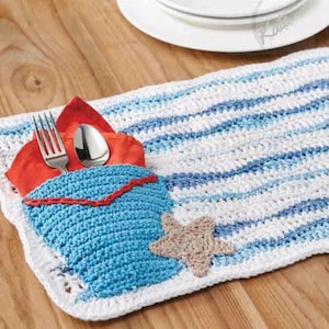 May include: A handmade crochet placemat with a blue pocket and a starfish accent. The placemat features blue and white stripes, a red napkin, and a silver fork and spoon. The pocket is trimmed with red yarn.