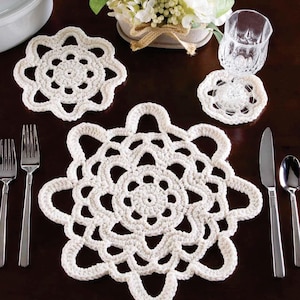 Op de afbeelding: Een verzameling handgemaakte, crèmekleurige gehaakte placemats en onderzetters, gerangschikt op een donkere houten tafel. De placemats hebben een bloemenpatroon en de onderzetters houden een glas vast. Zilverwerk en een klein bloemstuk zijn ook aanwezig.