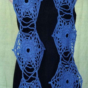 May include: A blue crochet scarf with a floral design. The scarf is displayed on a mannequin, showcasing its openwork pattern and central circular openings. The scarf's color is a vibrant blue.