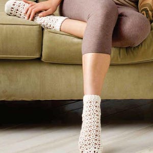 Pode incluir: Um par de meias de crochê brancas com um delicado padrão de renda. As meias estão sendo usadas por uma pessoa sentada em um sofá.