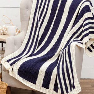 Puede incluir: Una manta a rayas azul marino y blanca sobre un sillón beige. La manta presenta gruesas rayas verticales y un borde de ganchillo. El plaid está hecho de un material suave y es perfecto para añadir calidez y estilo a cualquier espacio habitable.