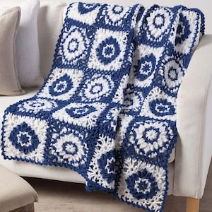 May include: A blue and white crocheted blanket draped over a white sofa. The blanket features a repeating pattern of square motifs with floral designs. The color scheme is a combination of navy blue and white, creating a visually appealing contrast.