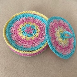 May include: A crocheted round box with a lid, featuring a multi-coloured design in shades of pink, blue, and yellow. The box has a small handle on the lid.