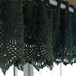 May include: Dark green crocheted curtain panels. The panels are made of a textured yarn with a scalloped edge. The crochet work has a detailed openwork design. The curtains are hanging on a white rod, with sunlight filtering through the openwork.