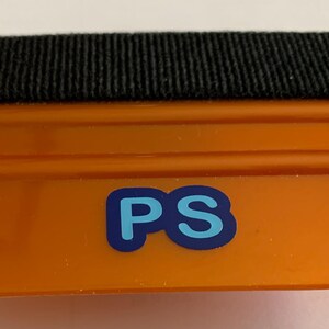 Può includere: Spatola in plastica arancione con bordo in feltro nero e logo "PS" blu.