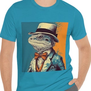 May include: A bright teal t-shirt featuring a graphic print of a lizard wearing a top hat, a suit jacket, and an orange bow tie. The lizard is depicted in a stylized manner against a split background of muted blue and orange tones.