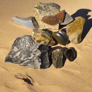 May include: A collection of various stones, including flint, quartzite, and jasper, arranged in a pile on a sandy surface. The stones are a mix of brown, gray, and green.