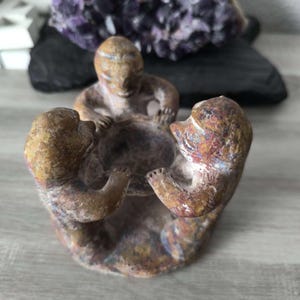 May include: A carved stone sculpture featuring three figures arranged in a circle, holding a central bowl-shaped basin. The sculpture has a mottled brown, tan, and reddish-orange texture, with smooth, rounded forms and defined facial features.
