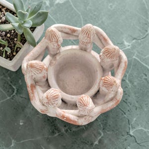 Peut inclure: Un bougeoir en céramique blanc et terracotta. Le bougeoir est conçu avec des figurines se tenant la main autour d'un bol central. Une plante succulente dans un pot blanc est en arrière-plan.