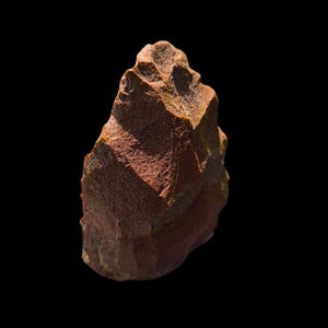 Pode incluir: Uma ferramenta de pedra castanho-avermelhada com uma superfície áspera e texturizada. A pedra tem uma borda lascada e uma ponta, sugerindo que foi usada para cortar ou raspar. O objeto está sobre um fundo preto.