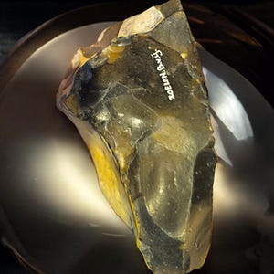 May include: A dark gray and yellow flint stone with the text "Zoelen Bay" inscribed on it. The stone is sitting on a dark brown plate.