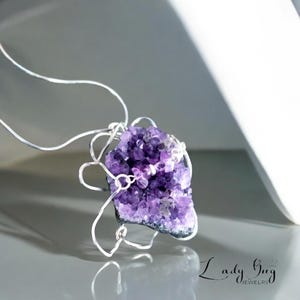 Op de afbeelding: Een zilveren ketting met een hanger van paarse amethistkristal. De kristal is gewikkeld in zilverdraad en heeft een natuurlijke, ruwe textuur. De hanger is versierd met kleinere amethistkristallen.