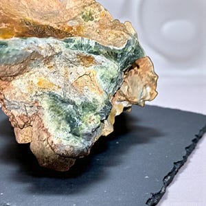 Puede incluir: Primer plano de una piedra natural rugosa con una mezcla de colores. La piedra presenta tonos marrones, verdes y beige, con texturas y patrones visibles. La piedra está colocada sobre una superficie oscura y rectangular.