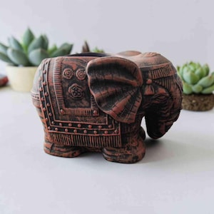 Op de afbeelding: Een terracotta-kleurige plantenbak in de vorm van een olifant met ingewikkelde reliëfpatronen aan de zijkant, waaronder cirkelvormige motieven en een dekenontwerp met franjes. Het beeld heeft een textuurafwerking met donkere schaduwen.