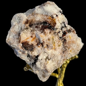 Könnte beinhalten: Ein weißer, grauer und brauner Kristallcluster mit einer rauen, strukturierten Oberfläche. Der Kristall befindet sich auf einem goldenen Metallständer.