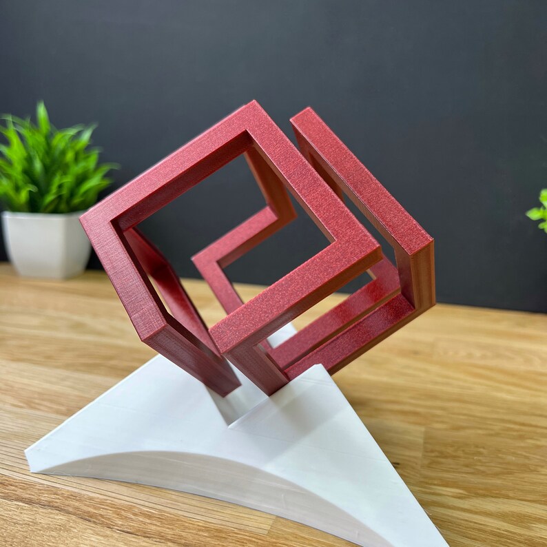 以下が含まれることがあります： 白い三角形の台座の上に置かれた、赤色の3Dプリントされた立方体の中に立方体がある彫刻。彫刻は、複雑で視覚的に興味深いデザインを生み出す、複数の層の連動する立方体で構成されています。
