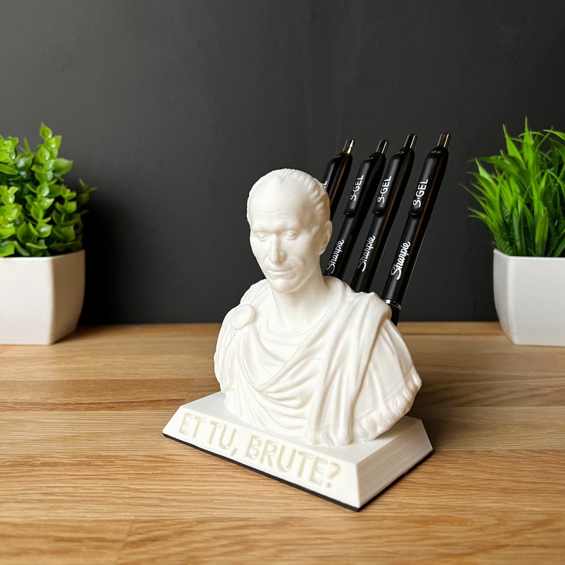 Puede incluir: Busto blanco de una figura romana con la inscripci&oacute;n "ET TU, BRUTE?" en la base. El busto se utiliza como portaplumas y tiene cuatro bol&iacute;grafos negros insertados en la parte posterior de la cabeza.