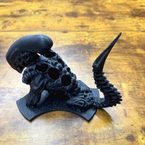 Puede incluir: Una figura de criatura alien&iacute;gena negra impresa en 3D con una cola larga y una cabeza que se abre para revelar una serie de agujeros. La figura est&aacute; colocada sobre una base negra.