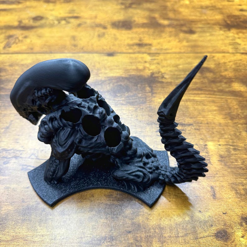 Puede incluir: Una figura de criatura alien&iacute;gena negra impresa en 3D con una cola larga y una cabeza que se abre para revelar una serie de agujeros. La figura est&aacute; colocada sobre una base negra.