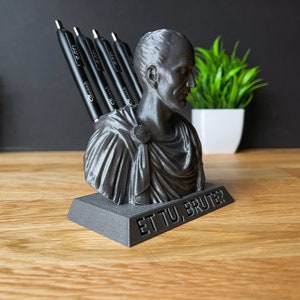 Puede incluir: Un porta l&aacute;pices de color negro y gris impreso en 3D con forma de busto de Julio C&eacute;sar. El porta l&aacute;pices tiene las palabras "Et Tu, Brute?" inscritas en la base.