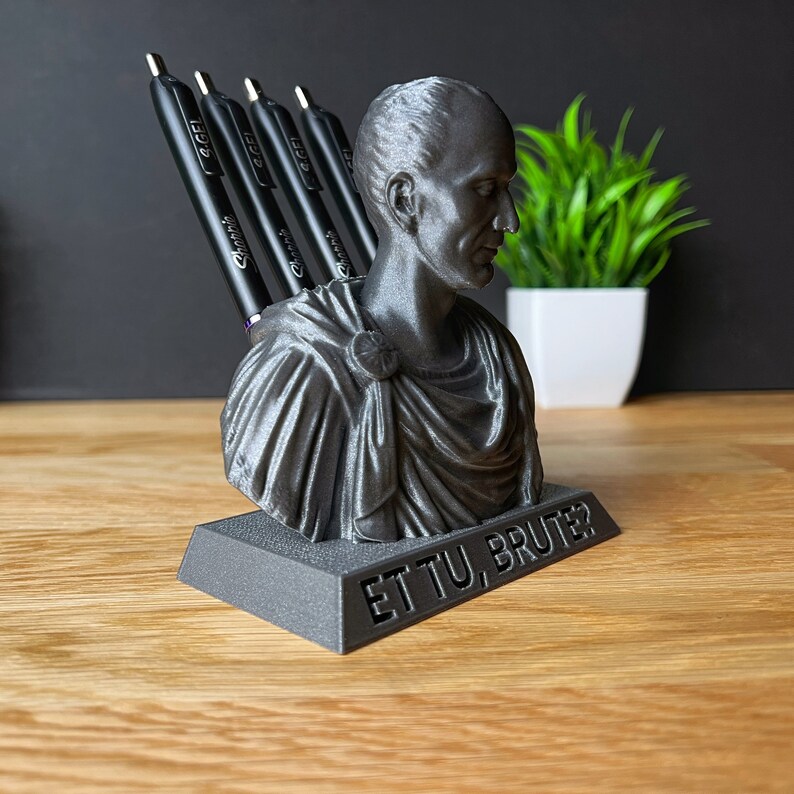 Puede incluir: Un porta l&aacute;pices de color negro y gris impreso en 3D con forma de busto de Julio C&eacute;sar. El porta l&aacute;pices tiene las palabras "Et Tu, Brute?" inscritas en la base.