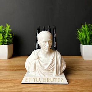 Puede incluir: Busto blanco de un emperador romano con la inscripci&oacute;n "Et Tu, Brute?" en la base. El busto se utiliza como portaplumas con cuatro plumas negras insertadas en la parte superior.