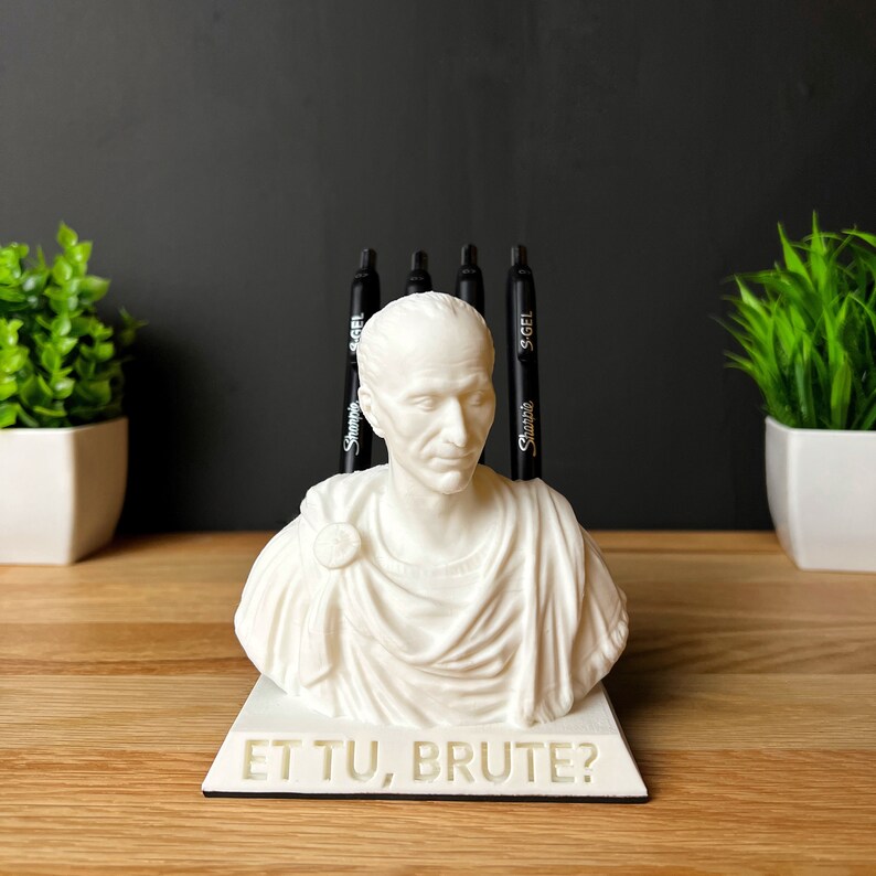 Puede incluir: Busto blanco de un emperador romano con la inscripci&oacute;n "Et Tu, Brute?" en la base. El busto se utiliza como portaplumas con cuatro plumas negras insertadas en la parte superior.