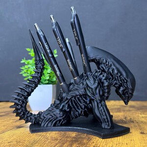 Puede incluir: Un portaplumas de criatura alien&iacute;gena negra impreso en 3D con cola. El soporte tiene cuatro bol&iacute;grafos negros insertados en &eacute;l. Los bol&iacute;grafos est&aacute;n etiquetados como "S-GEL" y "Sharpie".