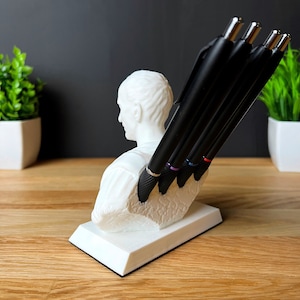 Puede incluir: Busto de escultura blanca de una persona con el pelo corto, utilizado como portaplumas. Cinco bol&iacute;grafos negros con detalles de color se insertan en el busto.