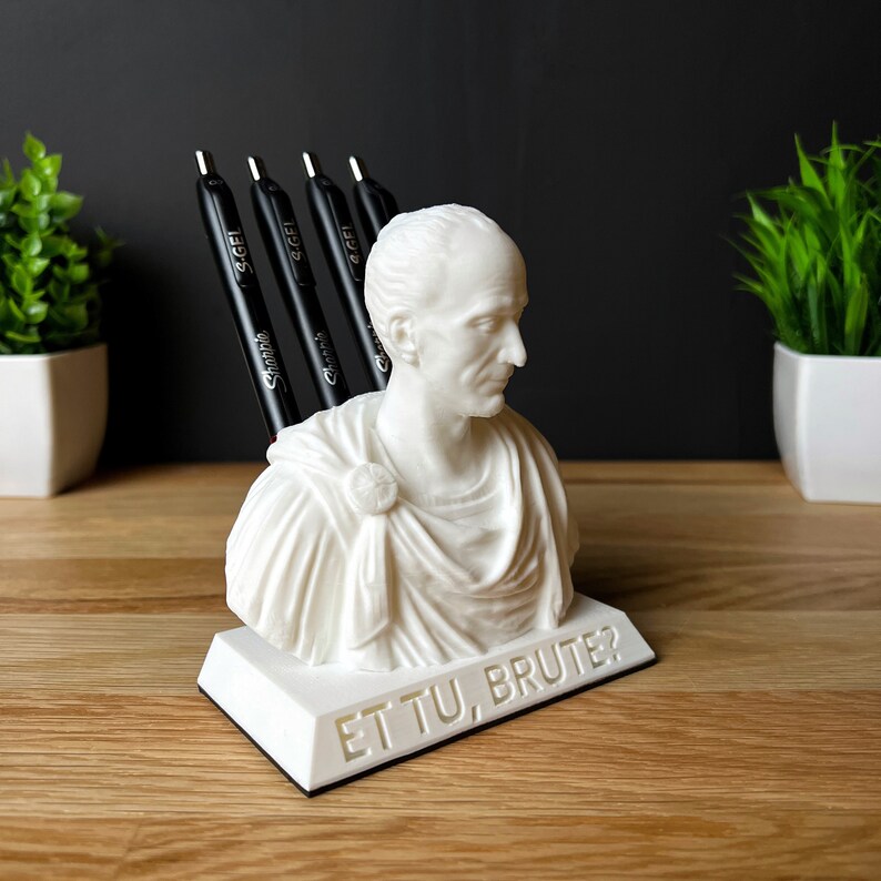 Puede incluir: Busto blanco de un hombre romano con toga, sosteniendo bol&iacute;grafos en la parte posterior. El busto est&aacute; sobre una base blanca con las palabras "Et Tu, Brute?" inscritas en la parte delantera.