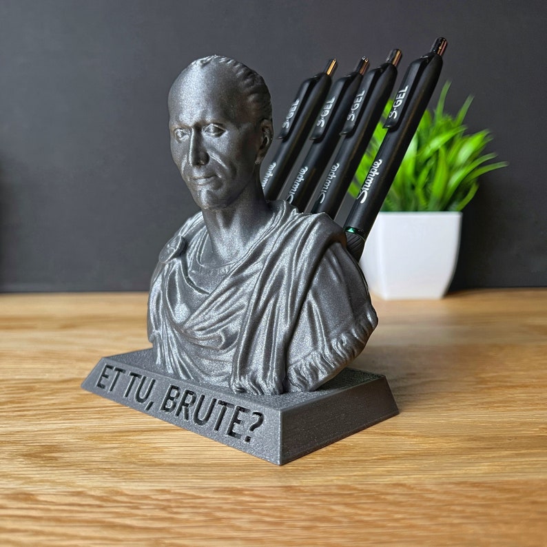 Puede incluir: Un organizador de escritorio impreso en 3D con forma de busto de Julio C&eacute;sar. El busto es gris y tiene la inscripci&oacute;n "ET TU, BRUTE?" en la base. El organizador contiene varios bol&iacute;grafos.