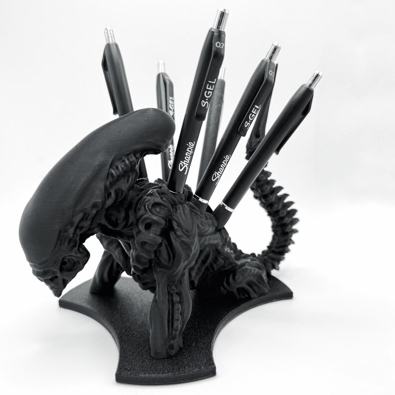 Puede incluir: Un porta-bol&iacute;grafos de criatura alien&iacute;gena negro impreso en 3D con cinco bol&iacute;grafos insertados. Los bol&iacute;grafos son negros y tienen las palabras "S-GEL" y "Sharpie" impresas en ellos.