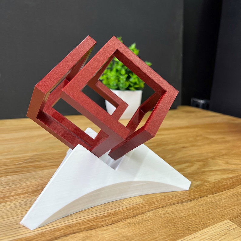 以下が含まれることがあります： 立方体の中に立方体が組み込まれた、赤い3Dプリントの幾何学的な彫刻。彫刻は白い湾曲した台座に載せられています。