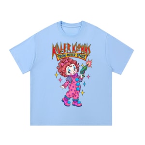 Puede incluir: Una camiseta azul claro con el gráfico de un payaso de dibujos animados con pelo rojo, que lleva un mono rosa con estampado de estrellas y botones azules, sosteniendo una pistola de rayos. El texto "KILLER KLOWNS FROM OUTER SPACE" está impreso en rojo y amarillo arriba.