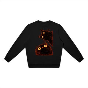 Puede incluir: Una sudadera negra de manga larga con un estampado gráfico de dos gatos negros con ojos amarillos grandes y abiertos y contornos naranjas. Los gatos están situados en el centro, uno mirando hacia abajo y el otro asomándose por un lado.